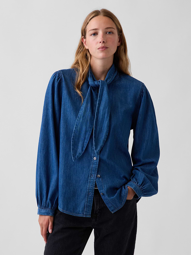 Image 1 of 5, Authentic, non-stretch denim button-up shirt., Tie neck, button front., Long sleeves with button cuffs., Shirring at back yoke seam., Shirttail hem., Medium wash., Responsibly Made: This denim shirt is part of our water-saving Washwell program. Compared with conventional wash methods, Washwell uses at least 20% less water., More Sustainable Cotton: Made with 55% Protocol cotton verified by the U.S. Cotton Trust Protocol, a program which works with farms committed to improving their environmental footprint by minimizing water usage and improving soil health (Claim No. 10127)., This product was made in a factory that invests in gender equity and women’s empowerment through RISE. Learn more at gapinc.com/equity ., Product #1184820