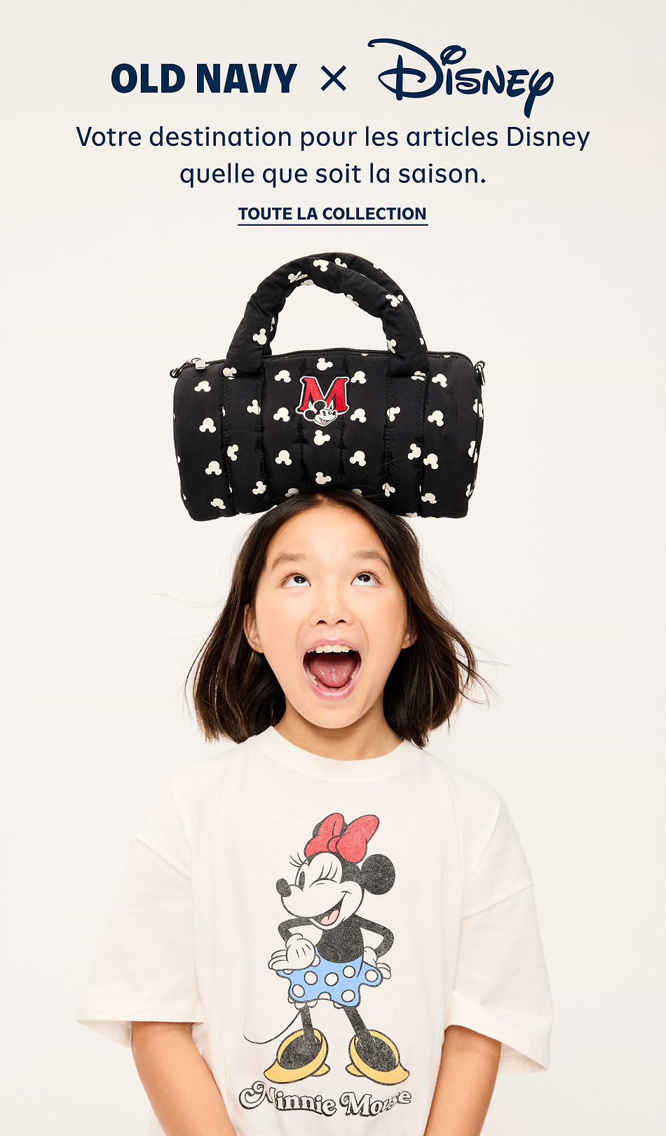 Child wearing a Minnie Mouse T-shirt balances a black Mickey-print duffel with a red “M” patch on their head; Old Navy x Disney ad.
