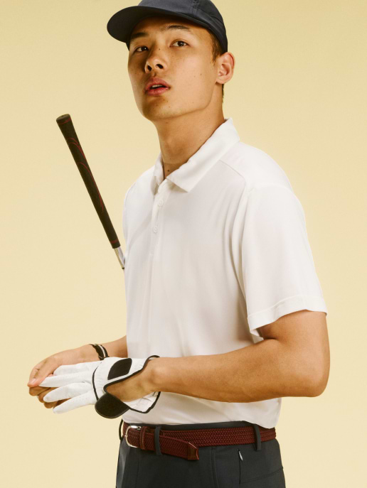 Golfer in a white polo and navy cap, club over shoulder, pulling on a glove against a beige background.