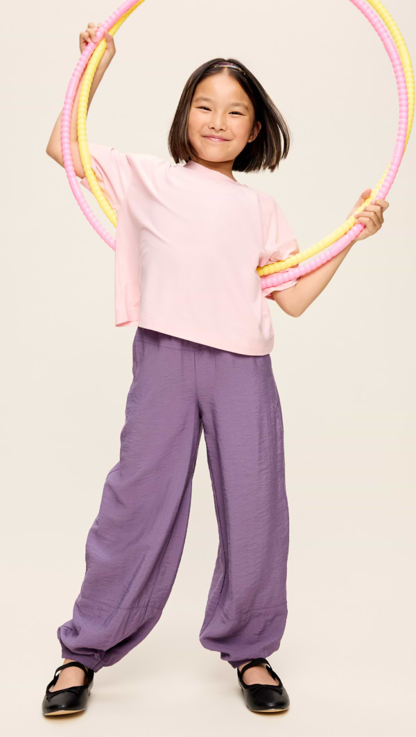 Smiling child stands holding a ring of pink and yellow hula hoops, wearing a light pink top, purple pants, and black flats.