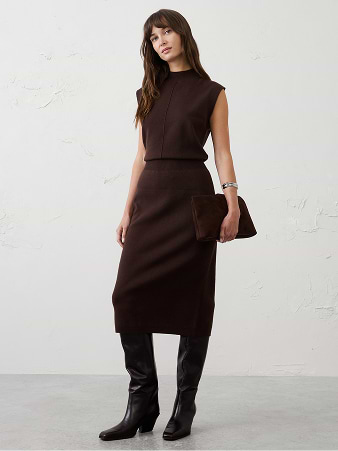Person in a dark brown sleeveless knit top and matching midi skirt, holding a clutch and wearing knee‑high boots.