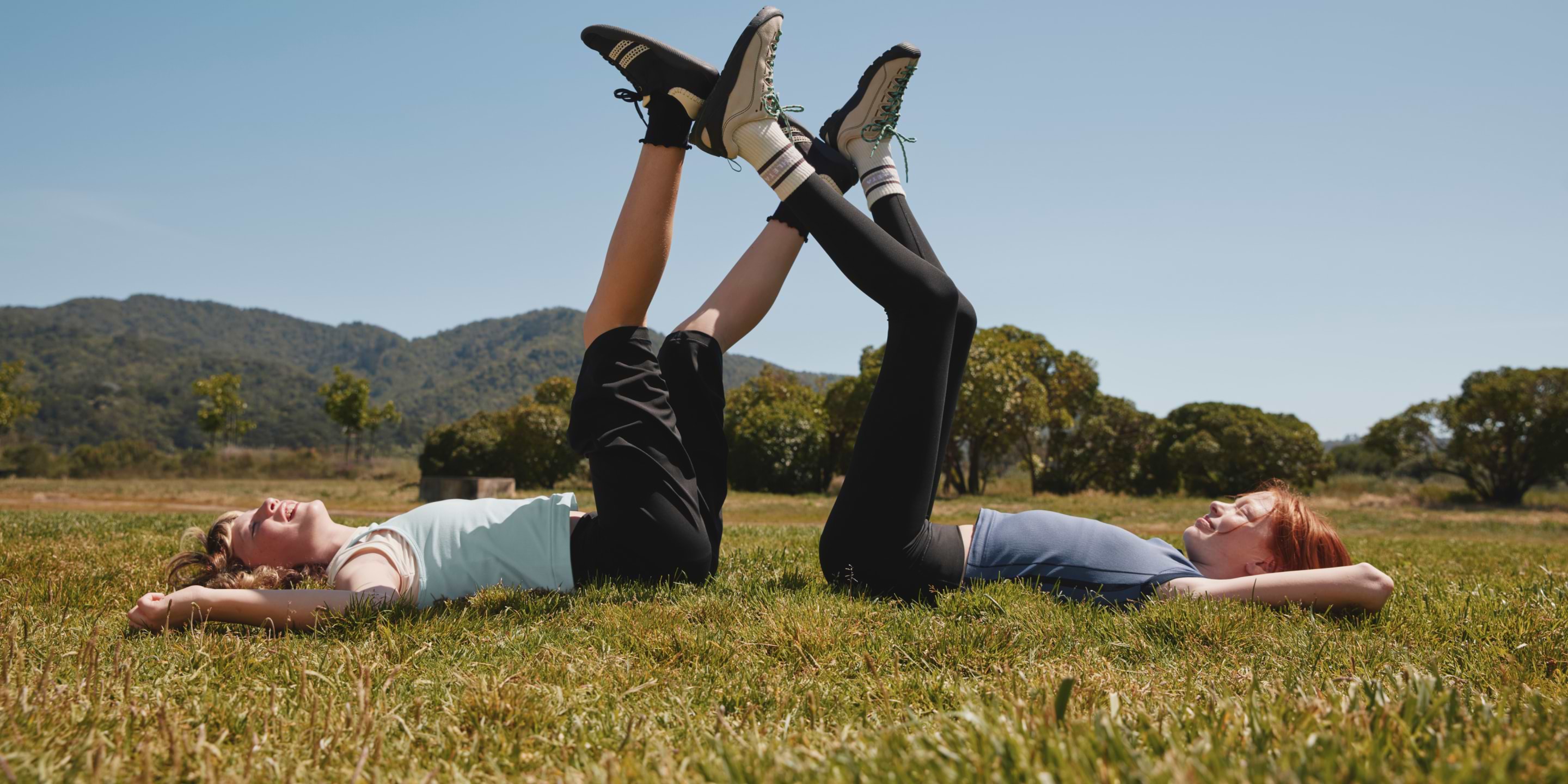 Two girls lie on a grassy field with legs raised and feet pressed together, smiling under a clear sky with hills and trees in the background.
