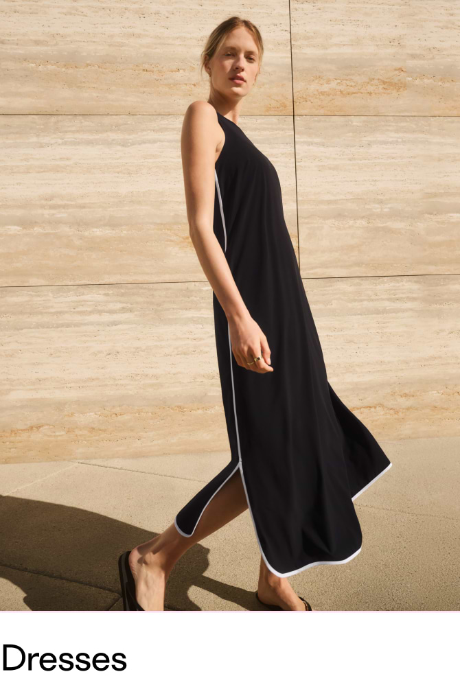Person walking outdoors in a sleeveless black maxi dress with white piping and side slits, wearing sandals by a tan stone wall.