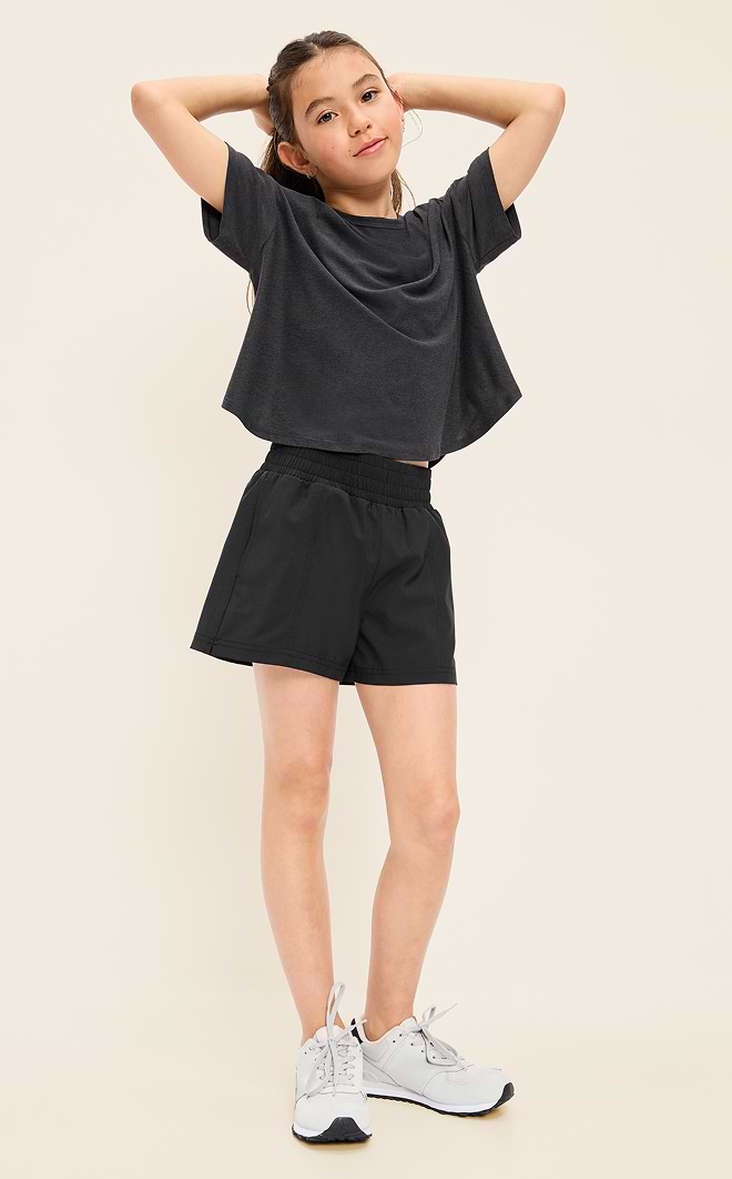 Child in a dark cropped T‑shirt, black athletic shorts, and white sneakers stands with hands behind head against a plain background.