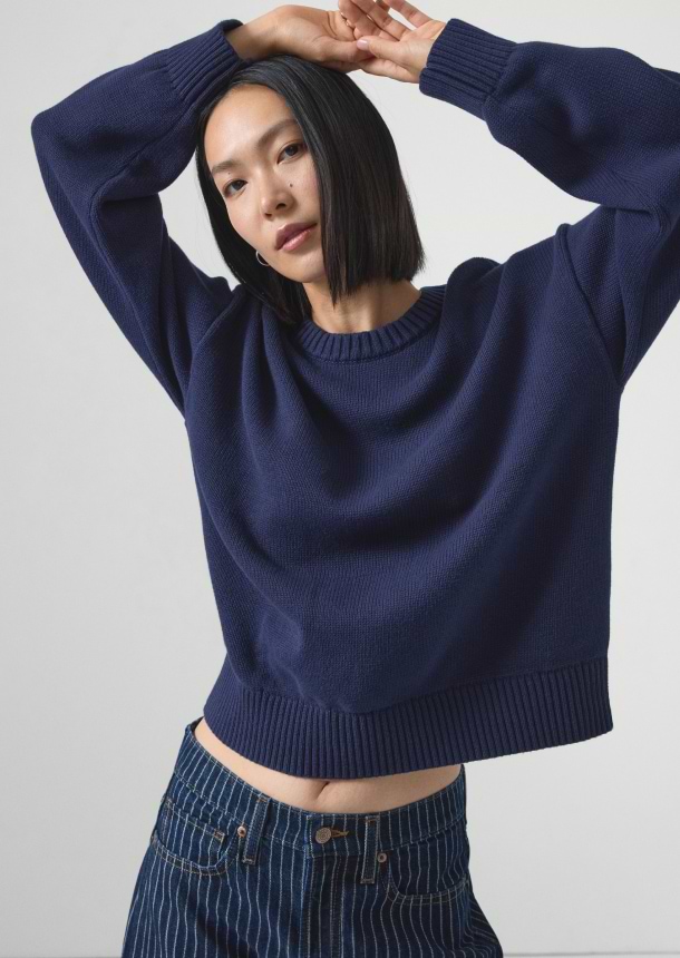 Person in a navy knit sweater and dark pinstripe jeans, arms raised above their head against a light background.