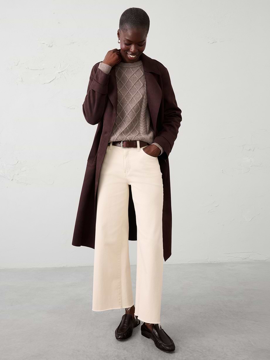 Smiling person wearing a dark coat, taupe sweater, cream wide-leg jeans, and brown loafers.