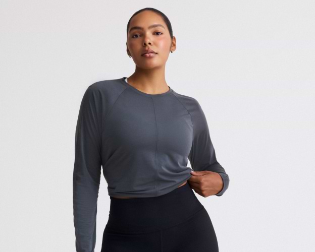 Person wearing a dark gray long-sleeve athletic top and black high-waisted leggings, holding the hem of the shirt against a plain background.