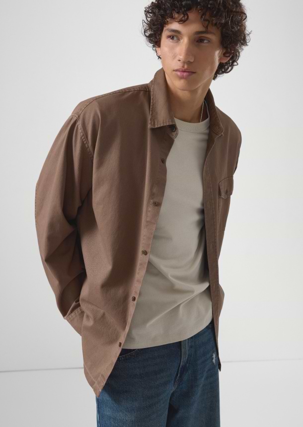 Person wearing a brown overshirt over a beige T‑shirt and blue jeans, looking sideways.