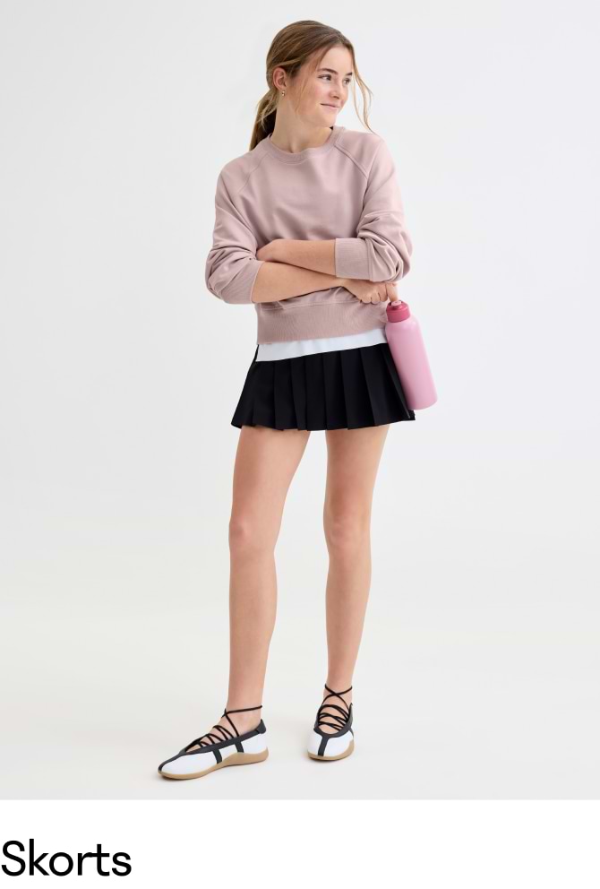 Girl in a pink sweatshirt and pleated black skort holds a pink water bottle, wearing black-and-white lace-up flats on a plain background.