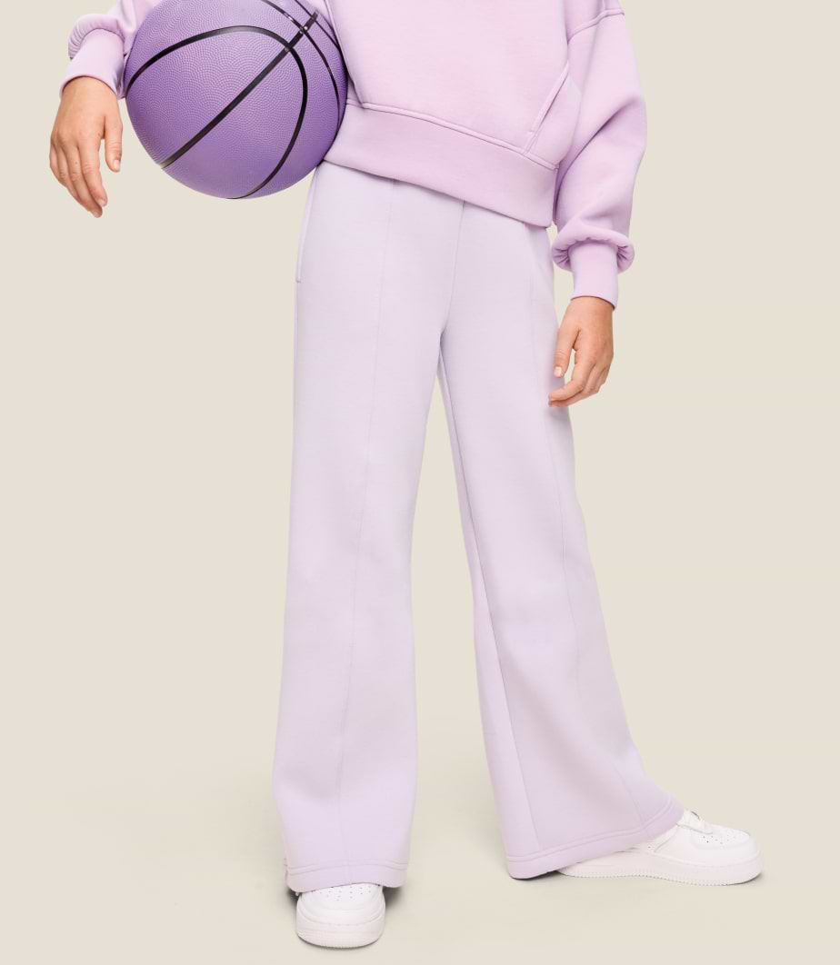 Cropped view of a person in lavender sweatshirt and wide-leg sweatpants holding a purple basketball, wearing white sneakers.