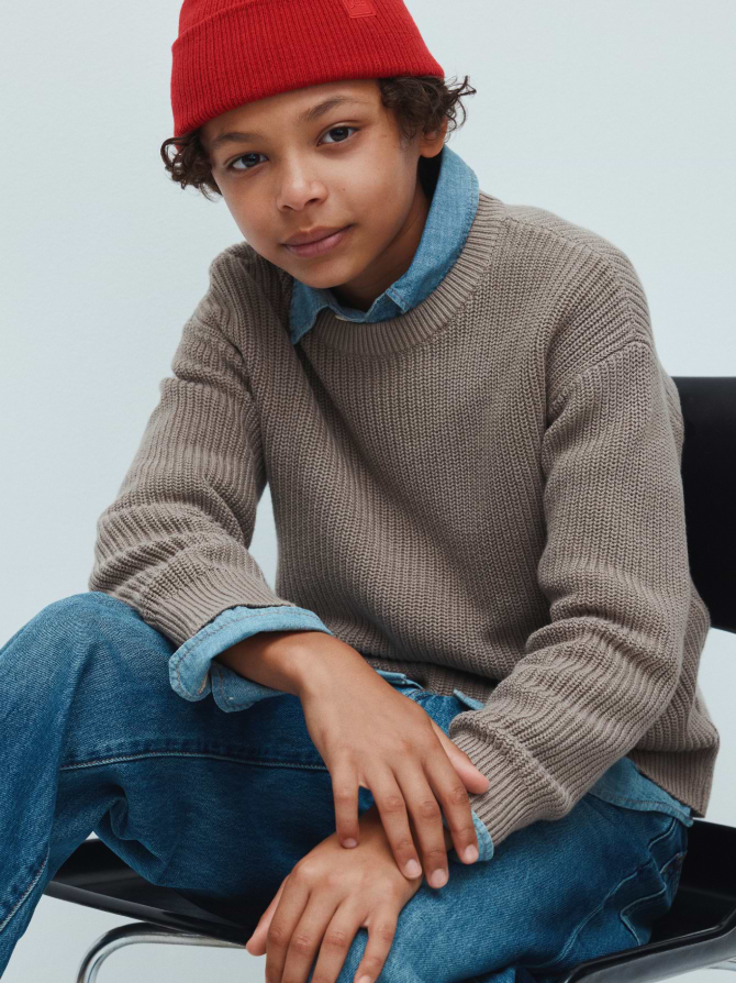 Young person in a red beanie, denim shirt and jeans under a tan knit sweater sits on a chair, looking at the camera.