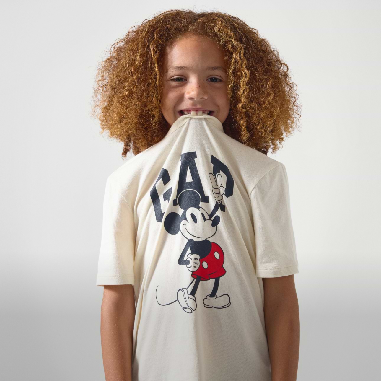 Smiling child biting the hem of a cream GAP T-shirt with a Mickey Mouse graphic.