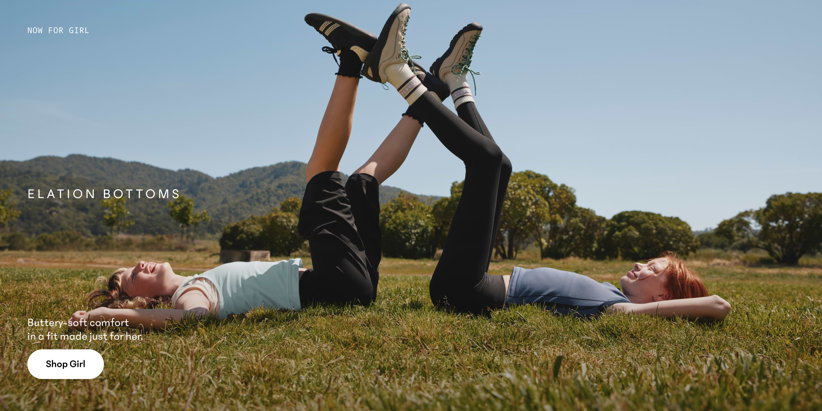 Two girls lie on grass in a sunny field, legs raised and feet touching, wearing leggings and sneakers with mountains in the background.