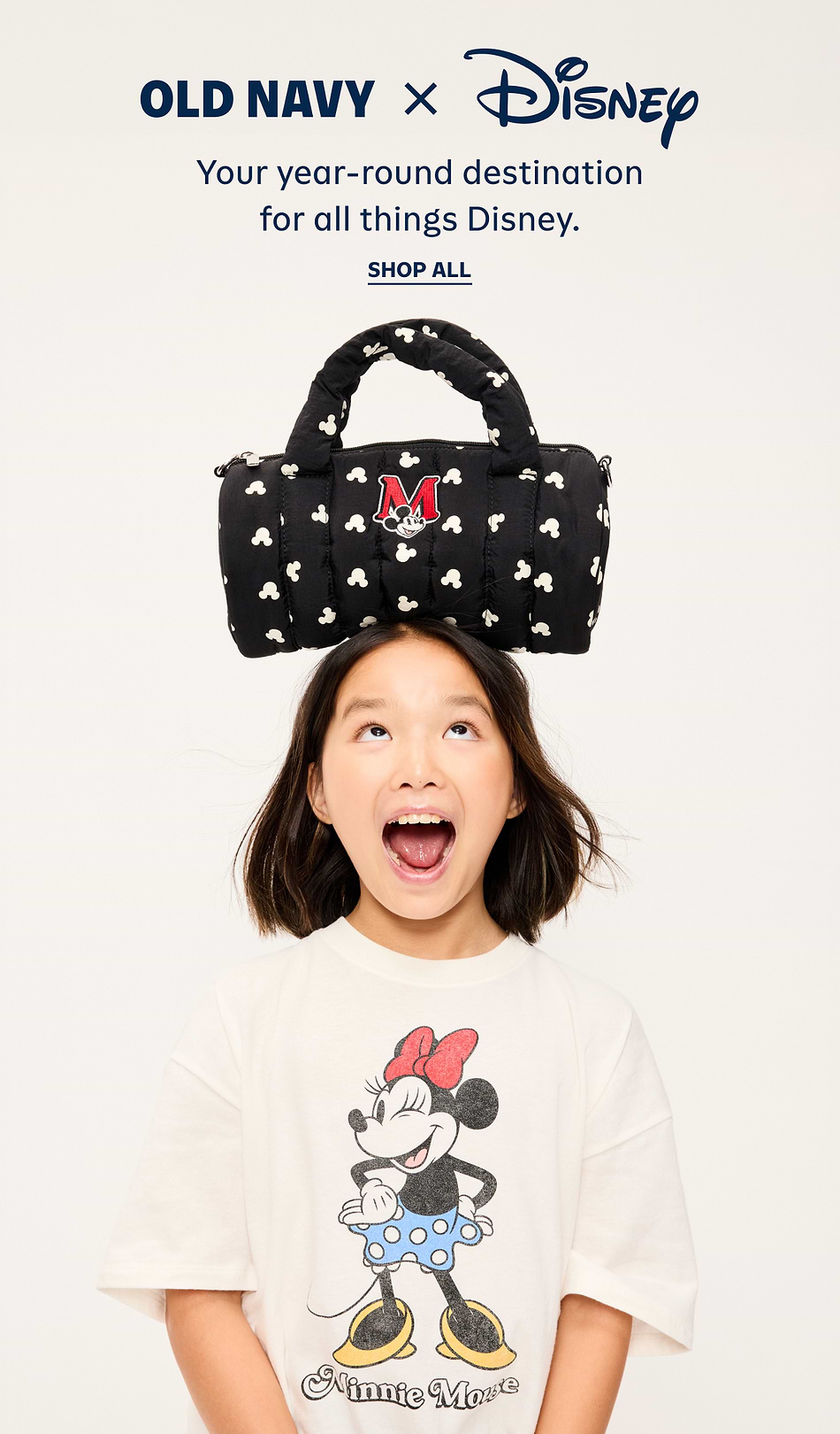 Child wearing a Minnie Mouse T-shirt balances a black Mickey-print duffel with a red “M” patch on their head; Old Navy x Disney ad.