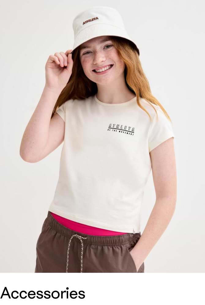 Girl wearing a cream Athleta logo T‑shirt and white bucket hat, hand on the brim, with brown drawstring pants and a pink layer showing, on a plain background.