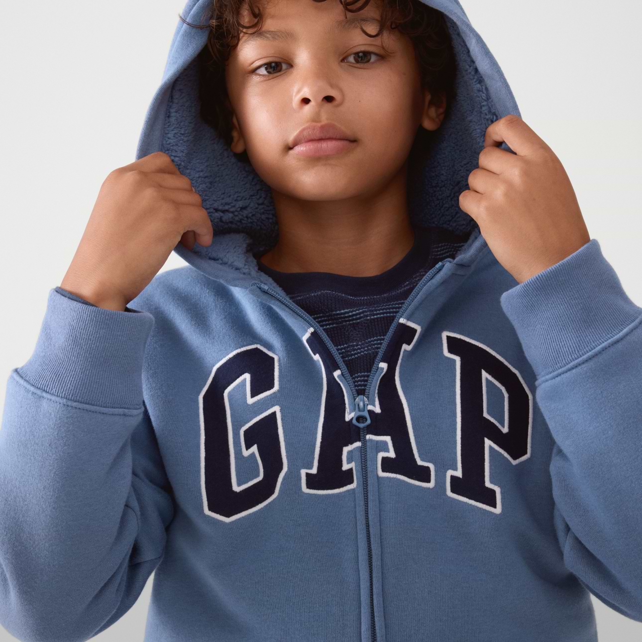 Child in a blue GAP zip-up hoodie, hood up, holding it.