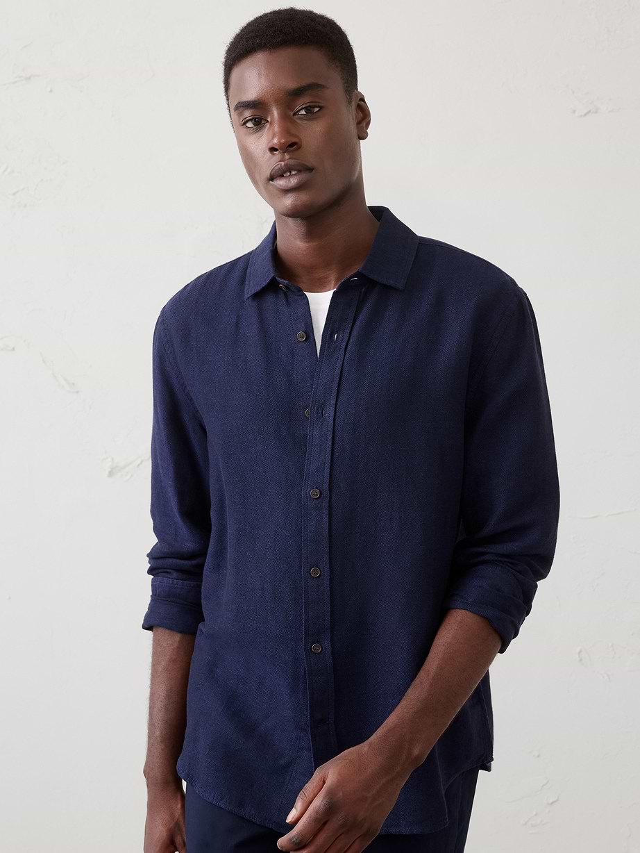 Person wearing a dark navy long-sleeve button-up shirt with rolled sleeves over a white tee, standing against a light wall.