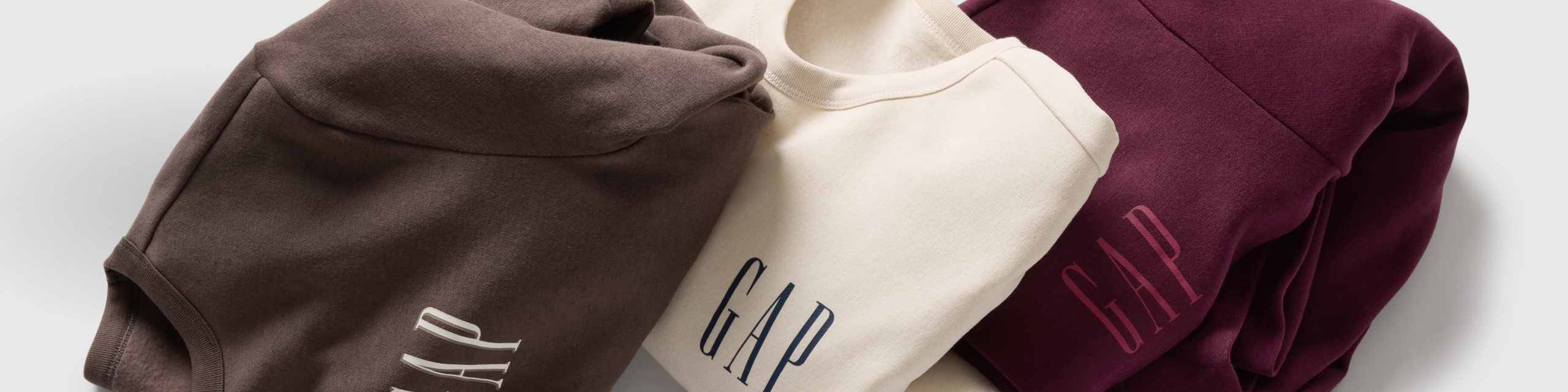 Three folded GAP sweatshirts: brown hoodie, cream crewneck, and burgundy hoodie.