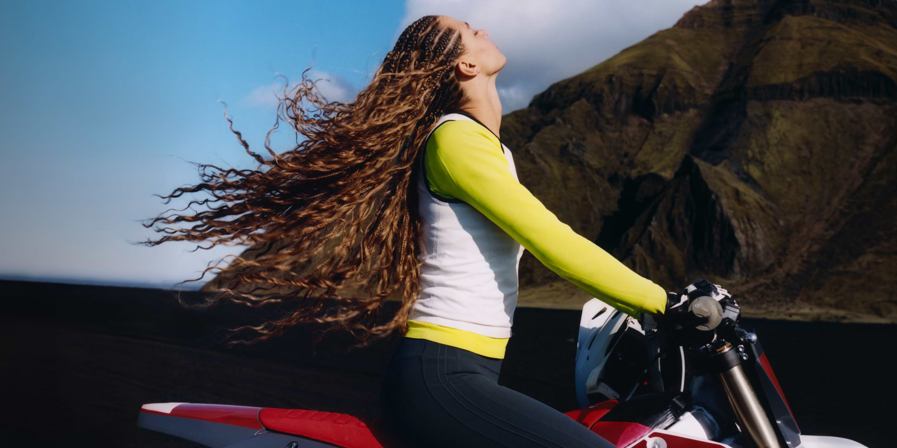 Person with long braided hair on a red dirt bike, head tilted back as hair flows, with rocky mountains and blue sky behind.