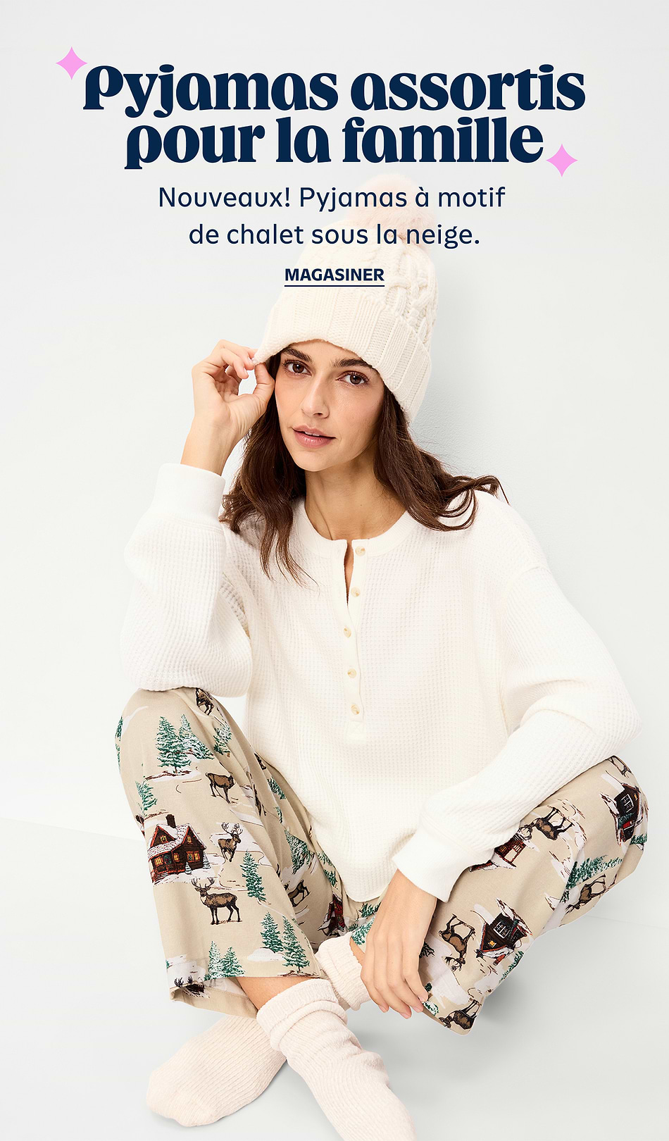 French ad for matching family pajamas: a woman in a cream knit top and beanie sits cross‑legged wearing beige pajama pants with snowy cabin and deer prints. Text reads: “Pyjamas assortis pour la famille – Nouveaux! Pyjamas à motif de chalet sous la neige. Magasiner.”