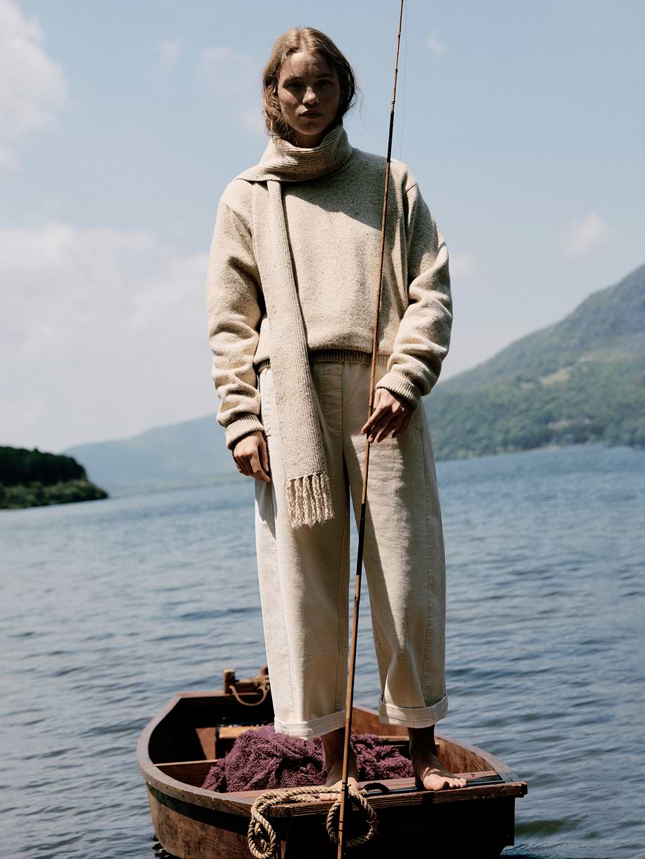Person in a beige sweater and scarf stands barefoot on a small wooden boat, holding a fishing rod on a calm lake with green mountains in the background.