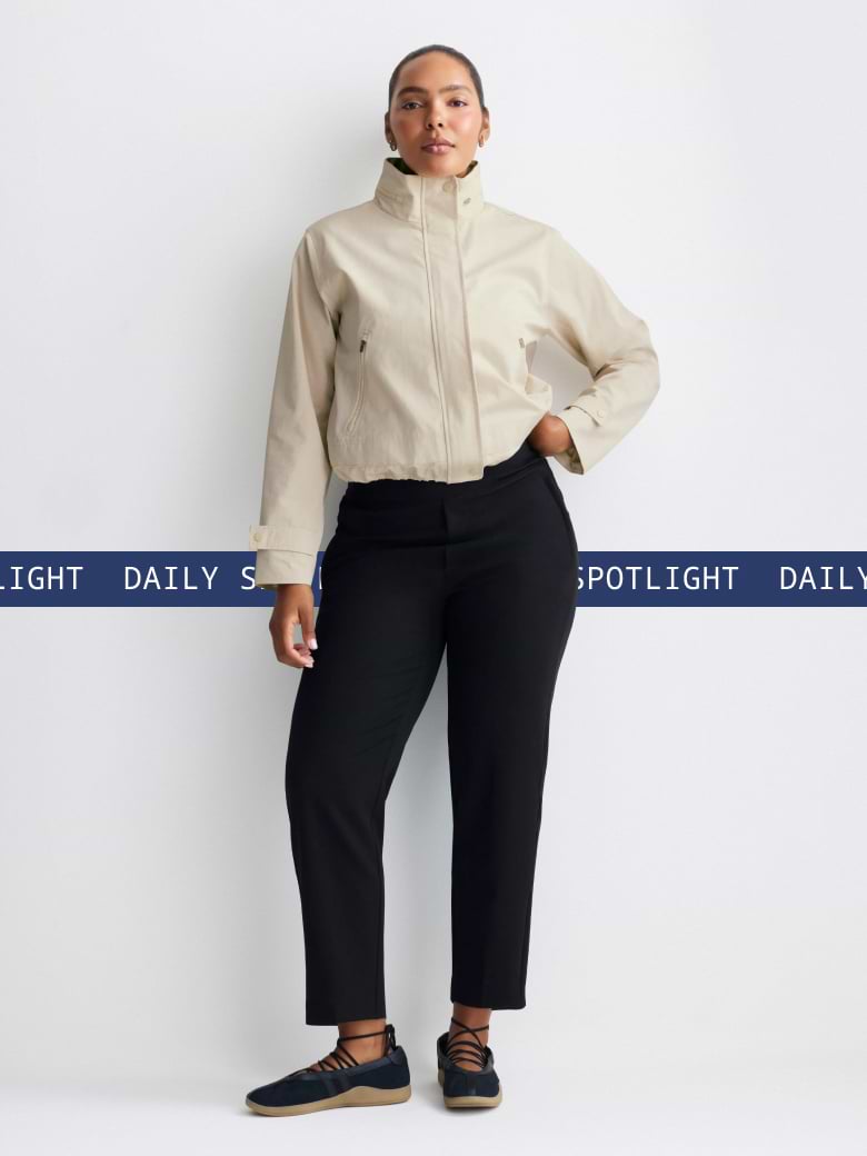 Model wearing a beige high-collar cropped jacket and black ankle pants with black lace-up flats, standing against a white background with a blue “DAILY SPOTLIGHT” strip across the mid-frame.