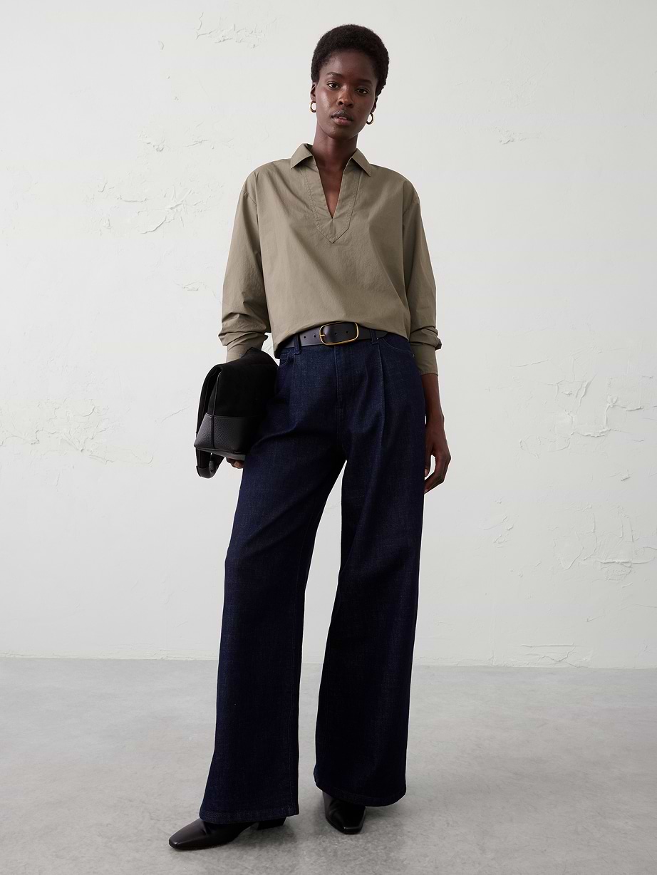 Person in an olive collared top and dark wide-leg jeans with a black belt and boots, holding a black bag.
