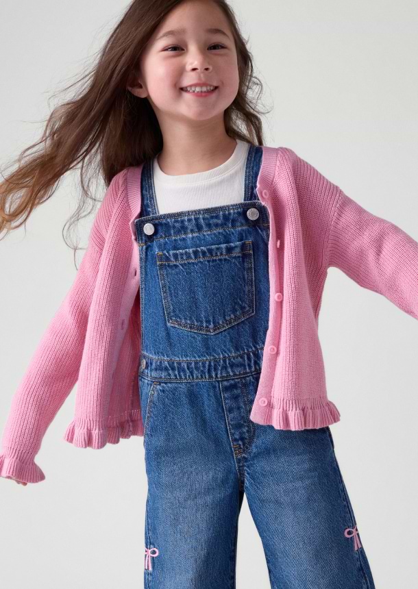 Smiling child in denim overalls and a pink ruffled cardigan, arms outstretched.