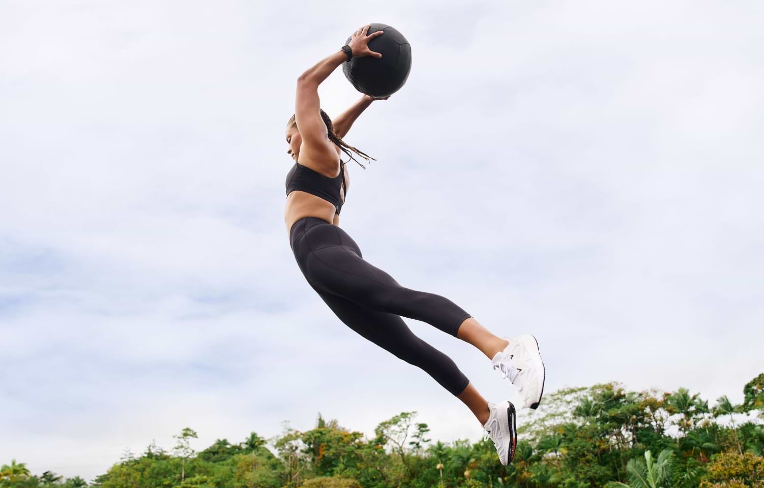 Woman wearing sunglasses and sporty attire strides across a rugged, rocky terrain. Green shrubs and distant hills populate the background under a partly cloudy sky.