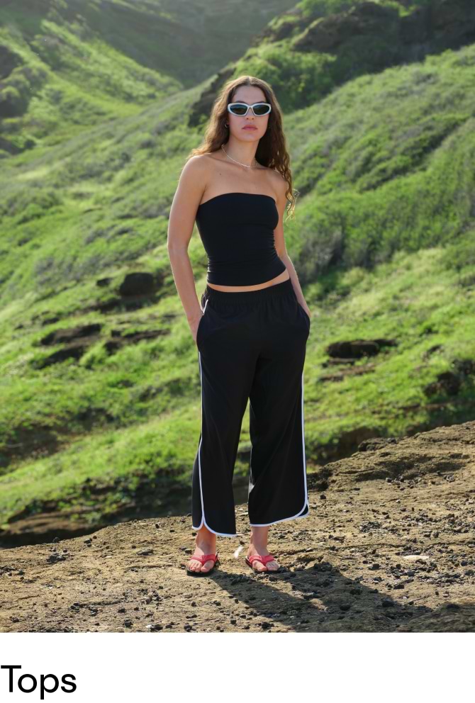 Person in a black strapless top and wide-leg pants, pink sandals, standing on a rocky hillside with green slopes in the background.