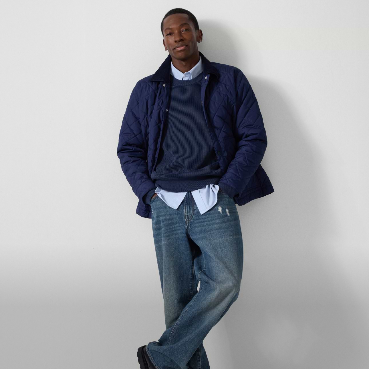Person leaning against a white wall, wearing a navy quilted jacket, blue sweater over a button-down shirt, relaxed blue jeans, and black boots.