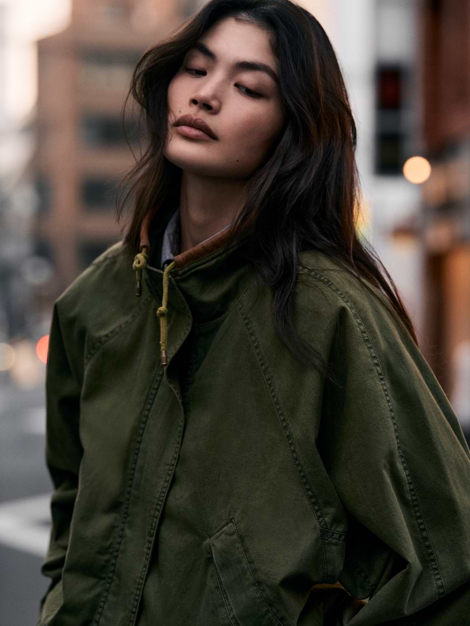 Person in an oversized olive-green jacket on a city street, looking down with a calm expression.