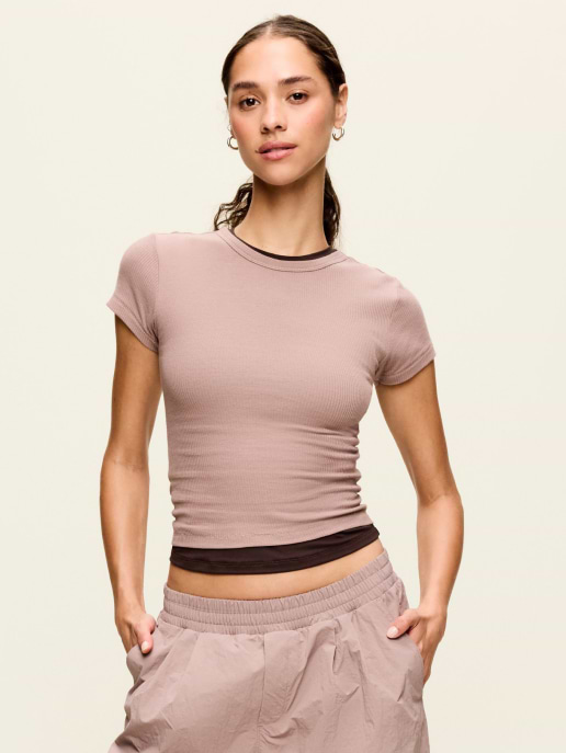 Person wearing a mauve ribbed fitted tee layered over a dark top and taupe joggers, hands in pockets, on a plain background.