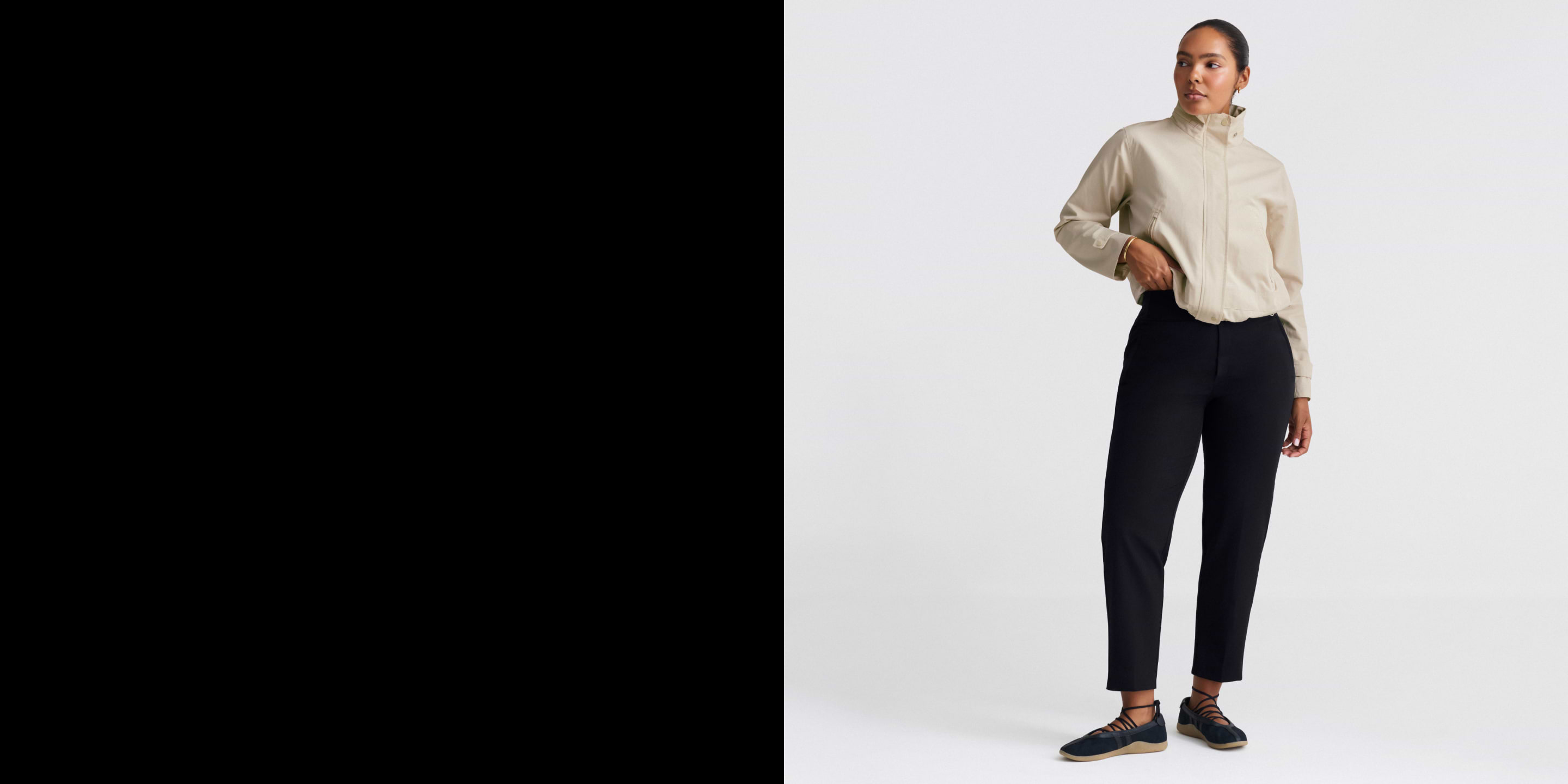 Person standing in a white studio wearing a beige high‑collar jacket, black ankle pants, and black lace-up shoes, one hand in a pocket.