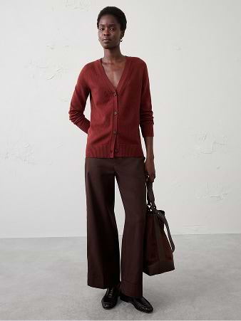 Person in a rust V‑neck cardigan and dark brown wide‑leg trousers, holding a brown tote bag, standing against a plain studio background.