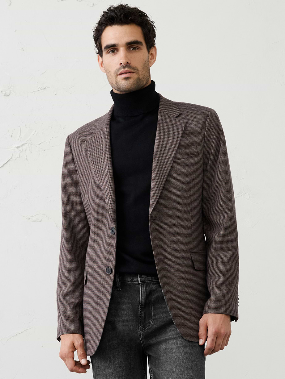 Person wearing an olive green diamond‑quilted jacket with dark brown collar and flapped chest pockets, standing against a plain light background.