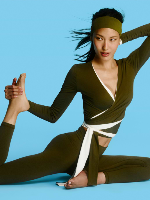 Person in olive green activewear does a seated stretch, holding one foot with a hand against a blue background.