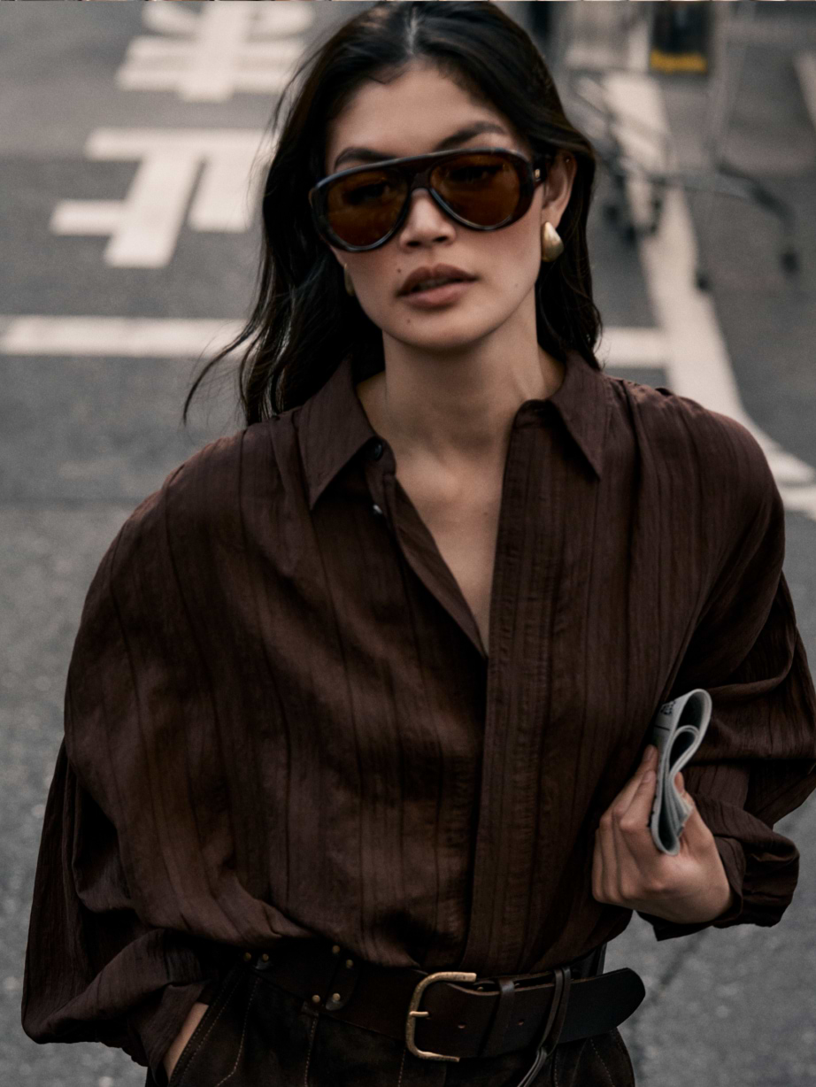 Woman in oversized sunglasses and a loose brown blouse walks on a city street, holding a rolled-up magazine.