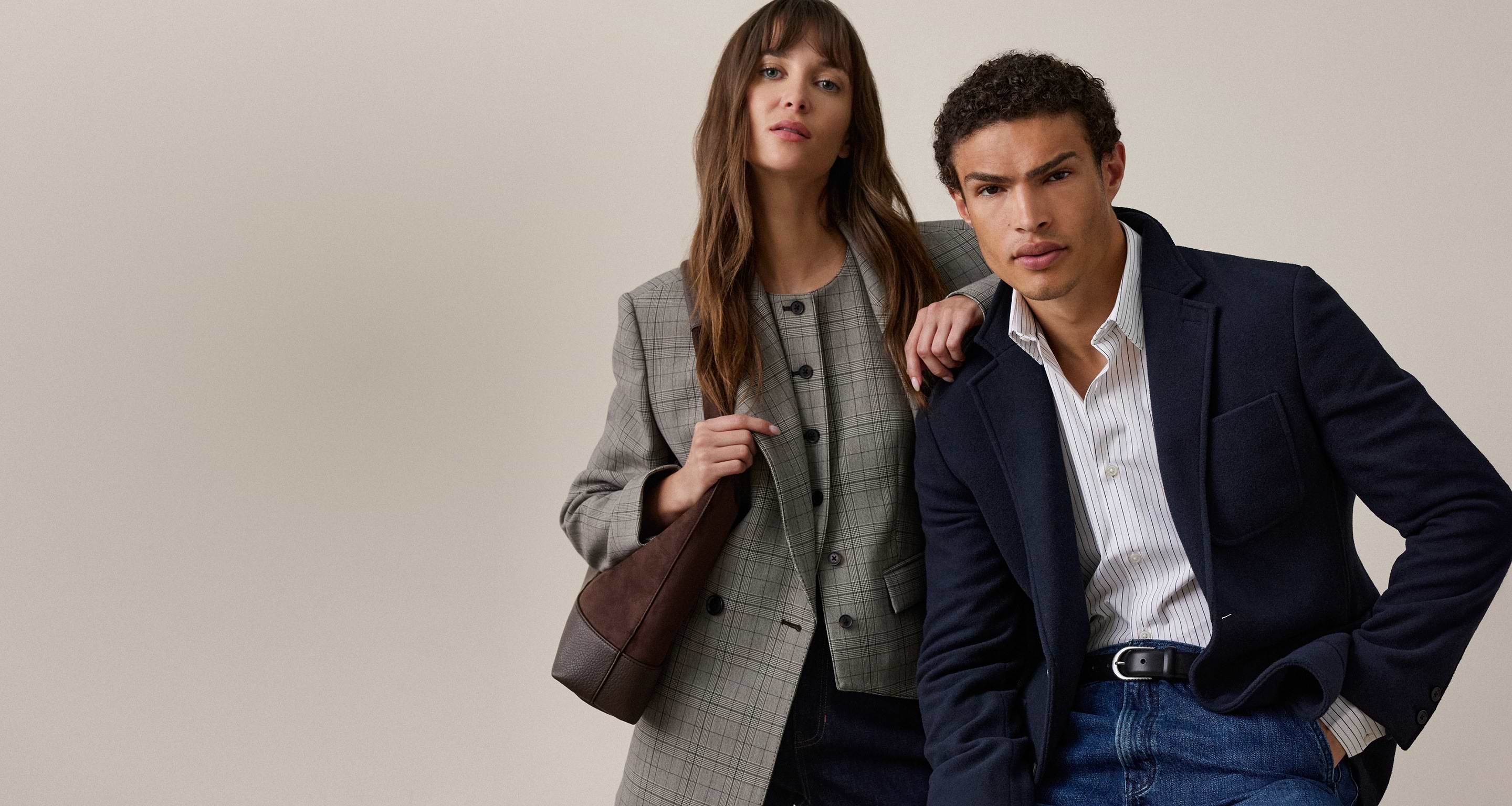 Two people in smart-casual outfits: woman in gray checked blazer with shoulder bag; man in navy blazer and striped shirt, against a plain backdrop.