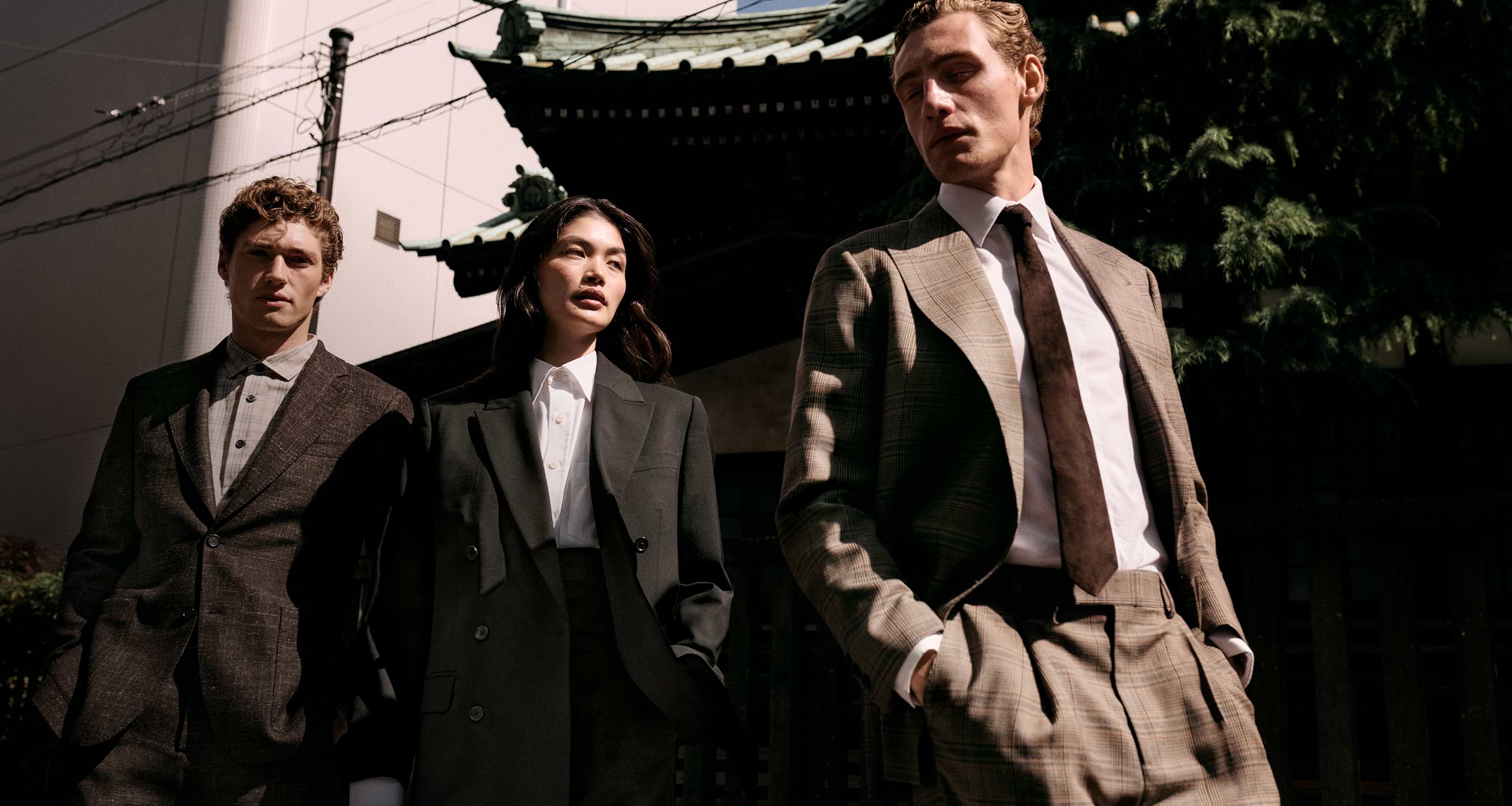 Three people in tailored suits walk past a traditional-style building on a sunny day.