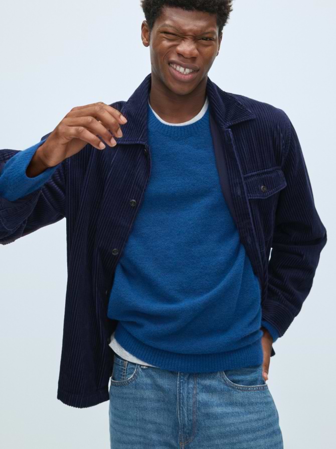 Person in a blue sweater and navy corduroy overshirt winks and raises a hand, standing against a plain light background.