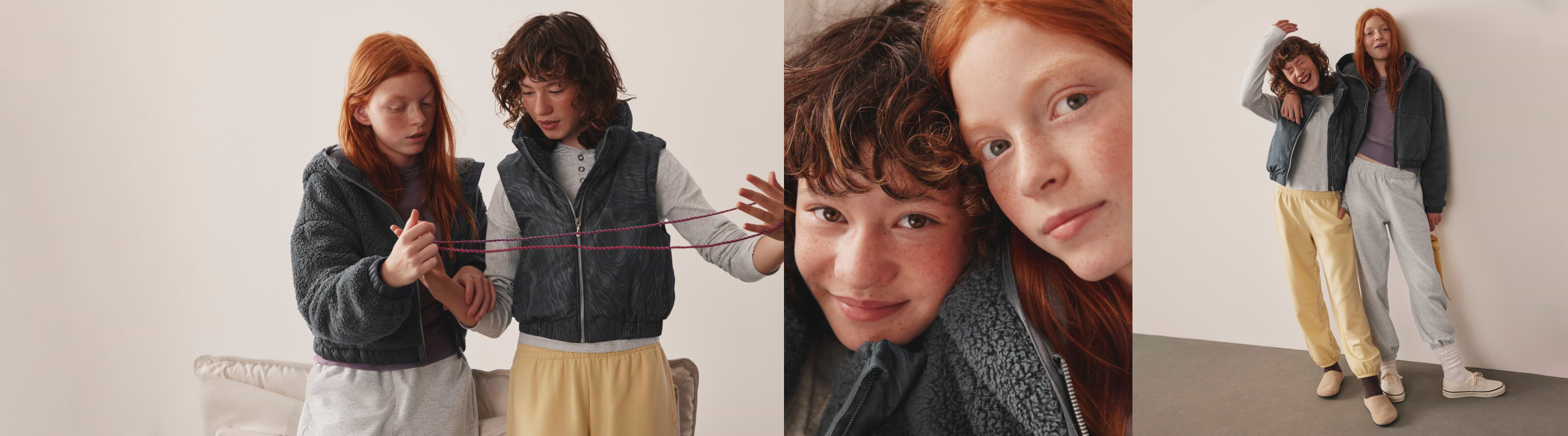 Two girls in cozy fleece layers and joggers play cat’s cradle, smile in a close-up, and stand arm-in-arm indoors.