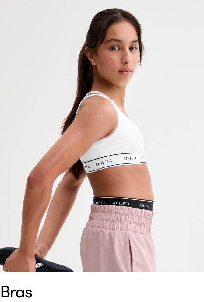 Side view of a Girl in a white Athleta sports bra and pink pants, holding shoes.