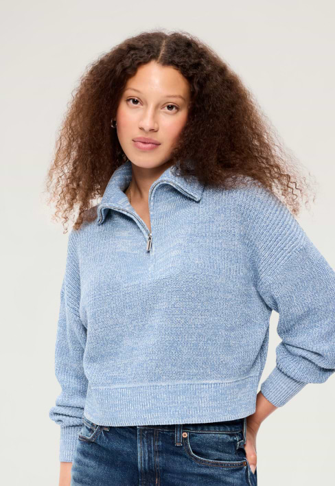 Person with curly hair wearing a light blue quarter-zip knit sweater and jeans against a plain background.