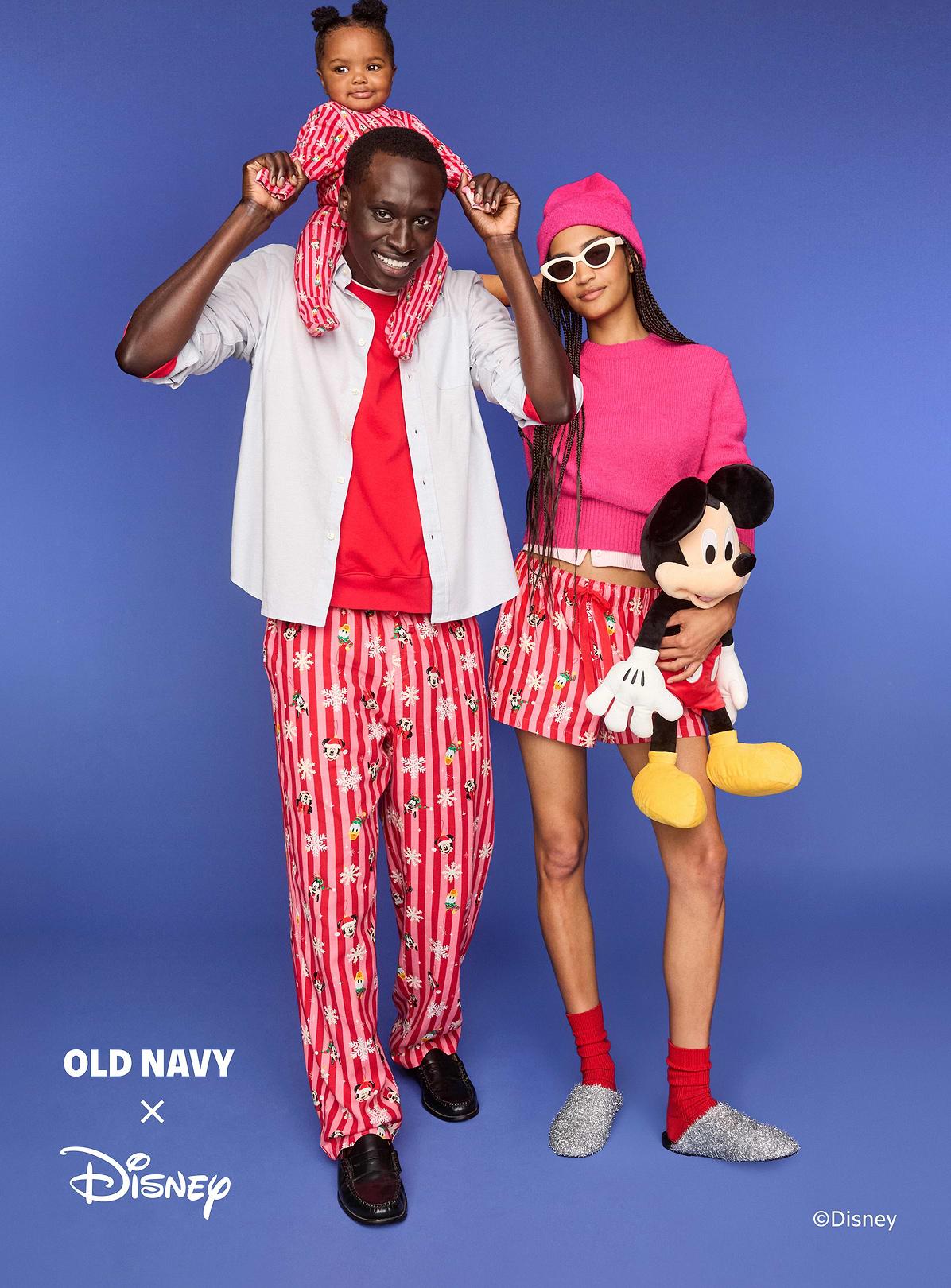 Two adults and a child in matching red, striped Disney pajamas pose on a blue background; the child sits on the man’s shoulders and the woman holds a Mickey Mouse plush. Old Navy x Disney logo visible.