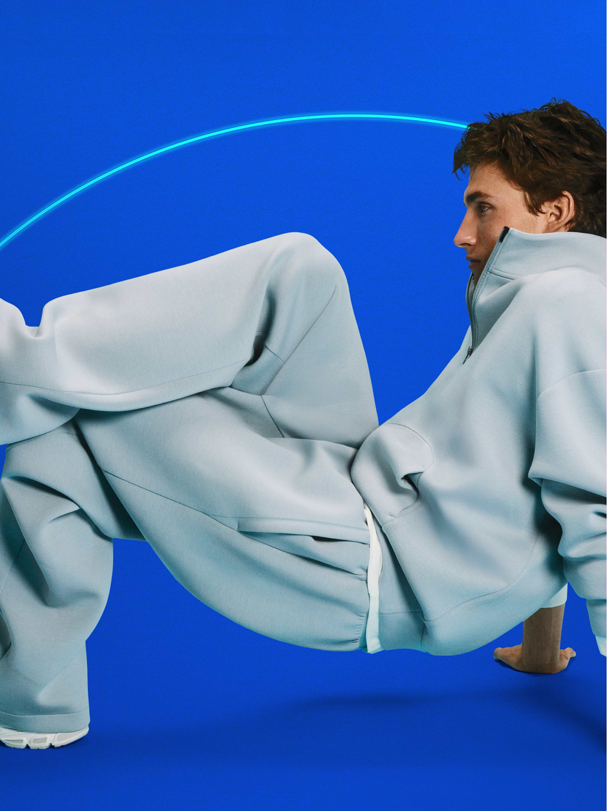 Person in oversized light-blue sweats balancing on hands against a bright blue background with a neon arc.