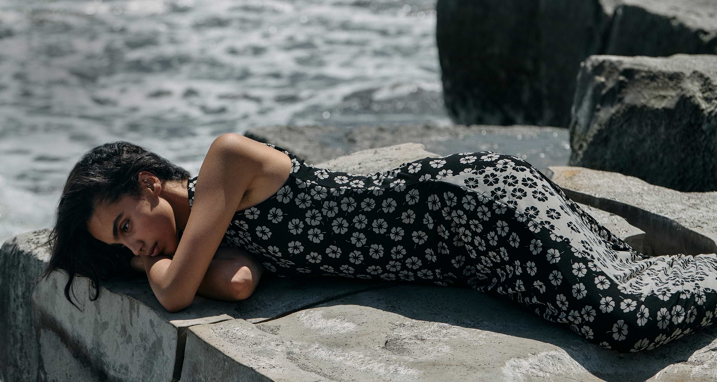 Person in a black floral dress lying on seaside rocks with waves behind.