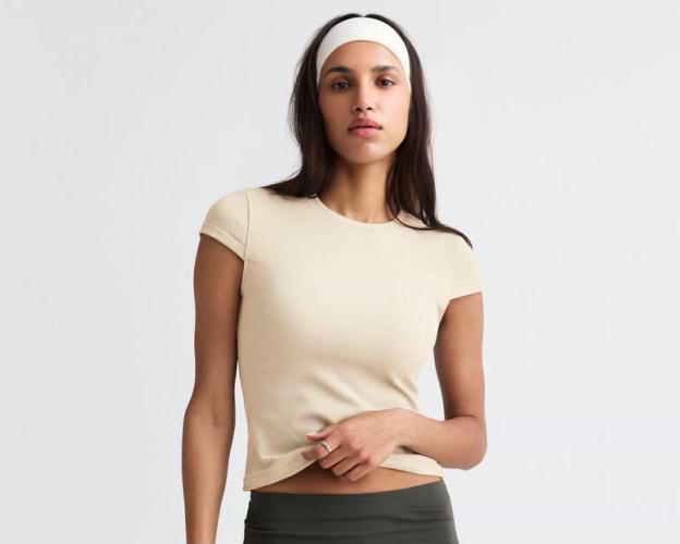 Person in a beige fitted T-shirt and white headband stands against a plain background, holding the shirt hem.