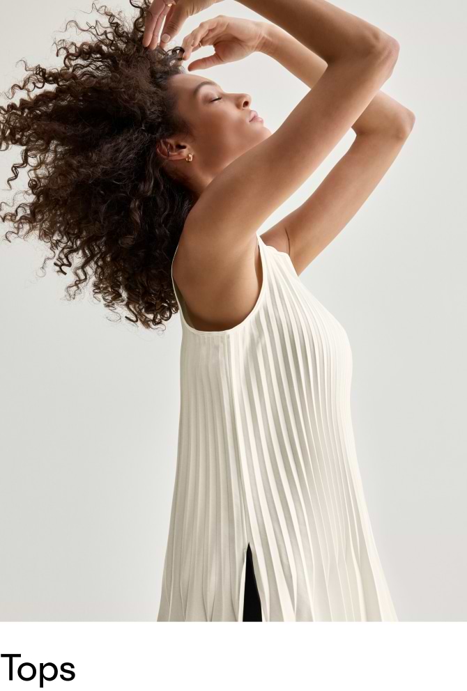 Woman with curly hair wearing a sleeveless, pleated ivory top with a side slit, arms raised.