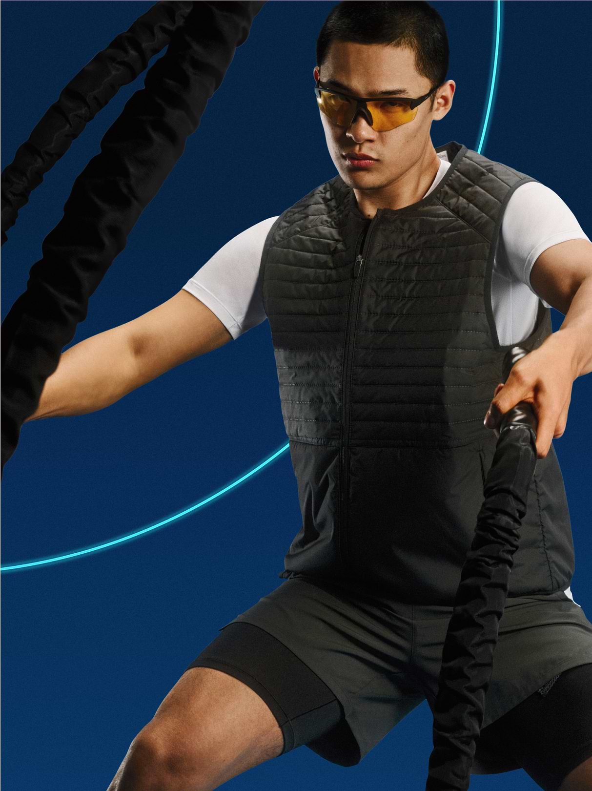 Athlete using battle ropes, wearing sunglasses, a quilted black vest, and shorts against a dark blue backdrop.